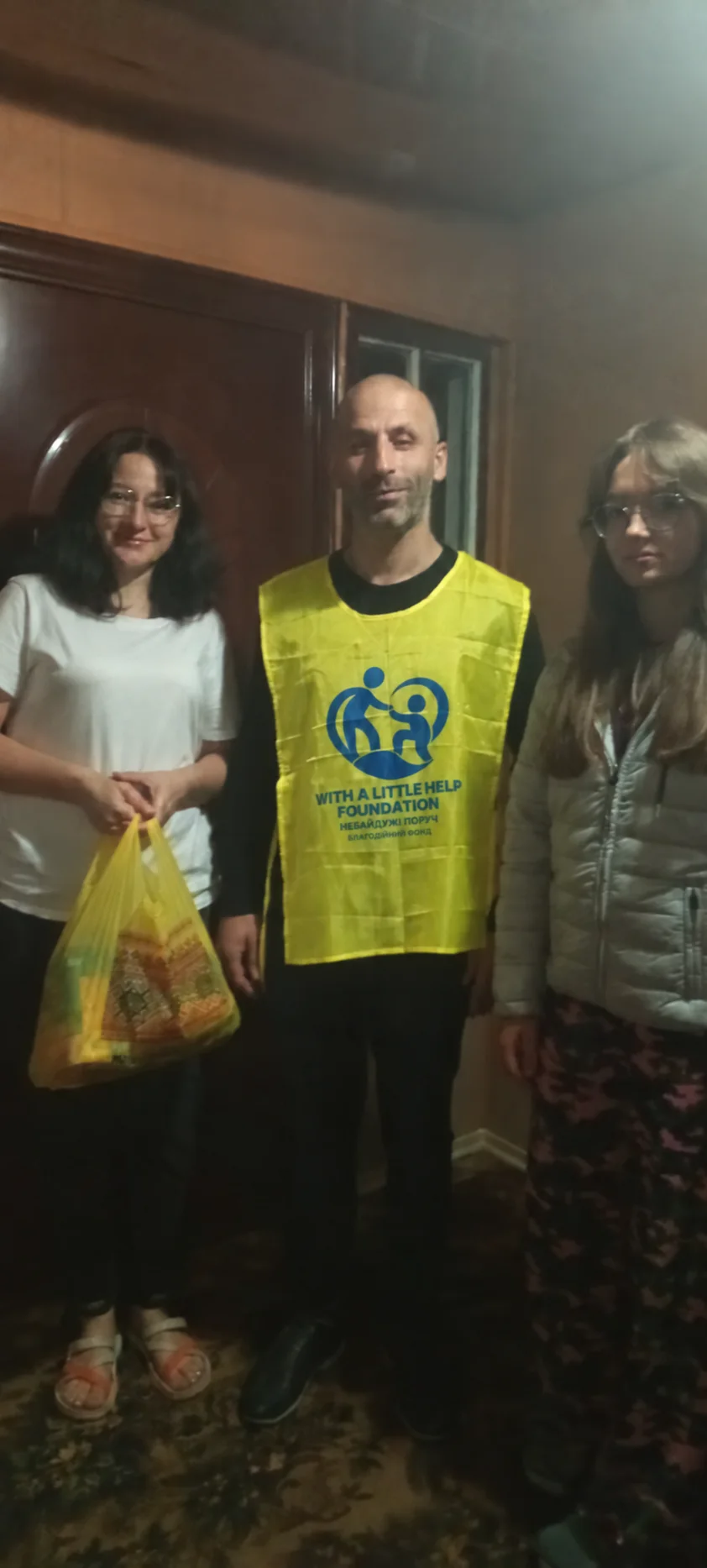 A team member standing indoors with two women, one holding a yellow bag.