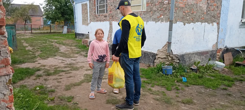 A team member seen from behind, wearing a yellow foundation vest and holding a bag of supplies beside children in a courtyard.