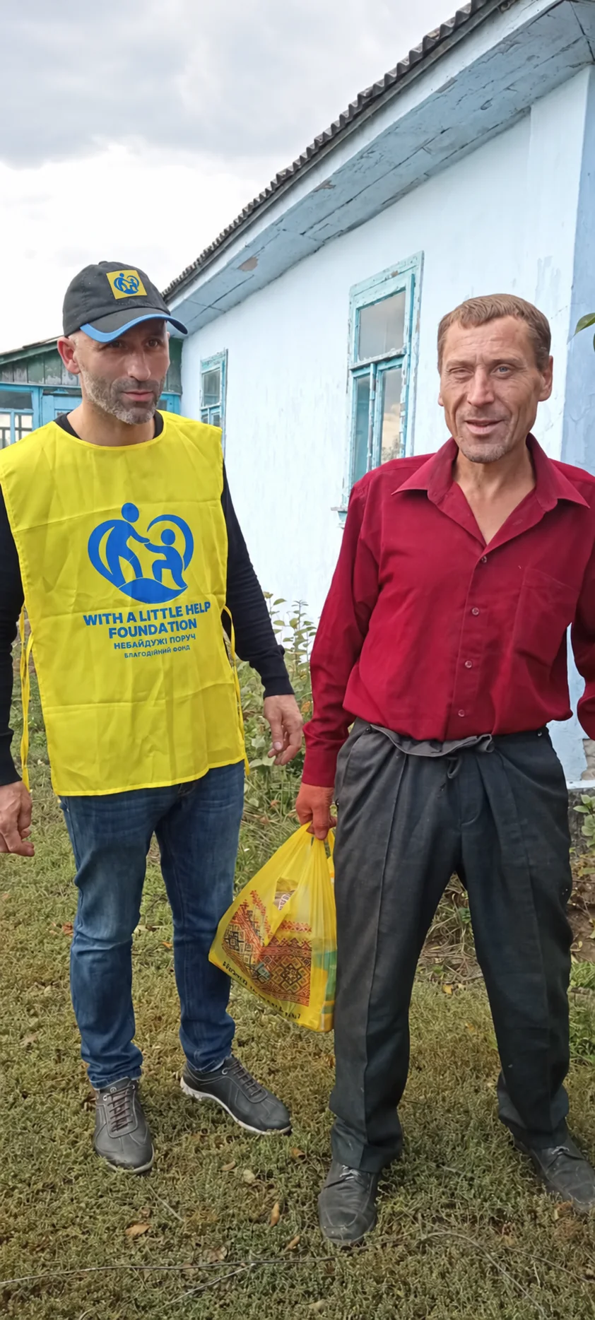 A team member and a man with a bag standing beside a blue building.